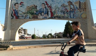 A young boy rides his bicycle in the southern Syrian city of Daraa on August 14, 2018. Behind him is a gate ornated with images of Syrian President Bashar al-Assad (L) and his late father Hafez al-Assad. (Photo by Andrei BORODULIN / AFP) (Photo credit should read ANDREI BORODULIN/AFP/Getty Images)